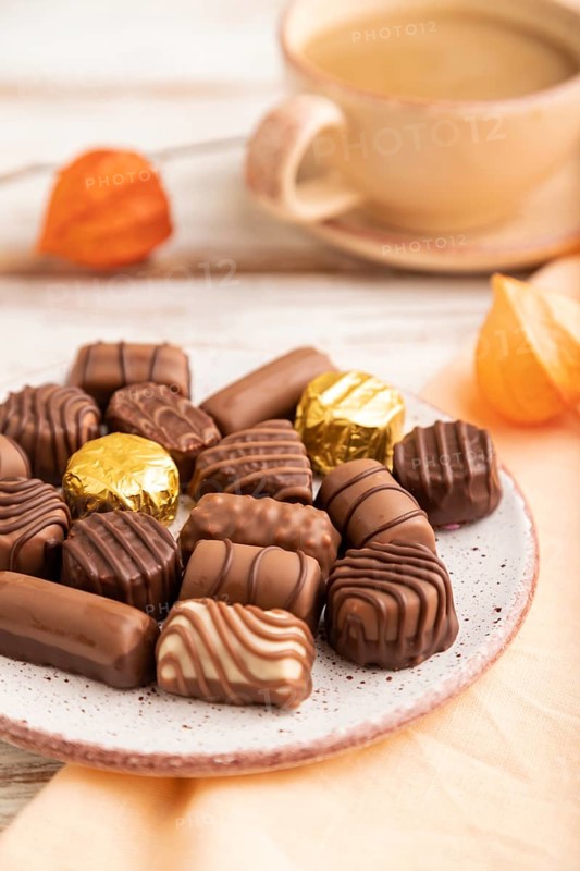 Chocolate candies with cup of coffee and physalis flowers on a white wooden background and orange textile. side view, close up, selective focus