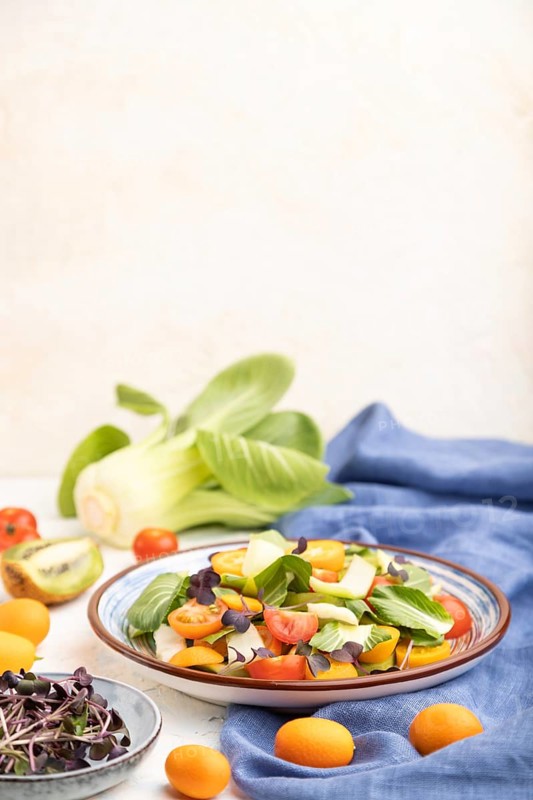Vegetarian salad of pac choi cabbage, kiwi, tomatoes, kumquat, microgreen sprouts on a white concrete background and blue linen textile. Side view, selective focus, copy space