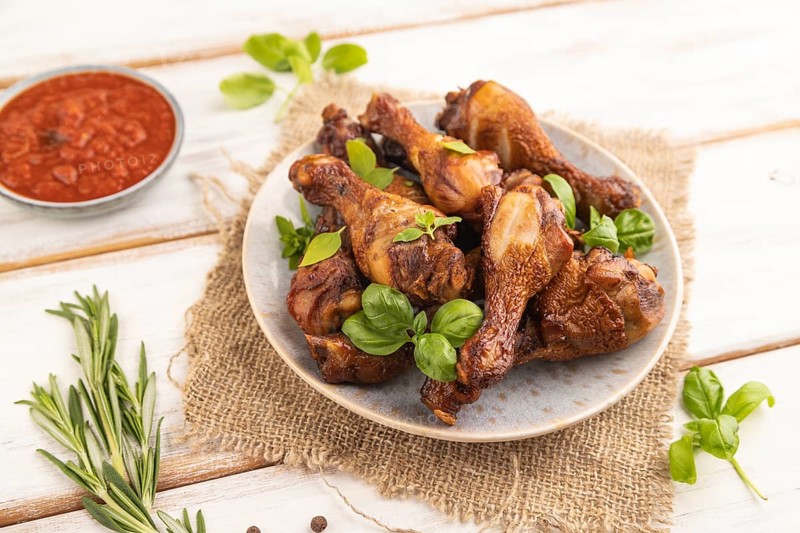 Smoked chicken legs with herbs and spices on a ceramic plate with linen textile on a white wooden background. Side view, close up, selective focus