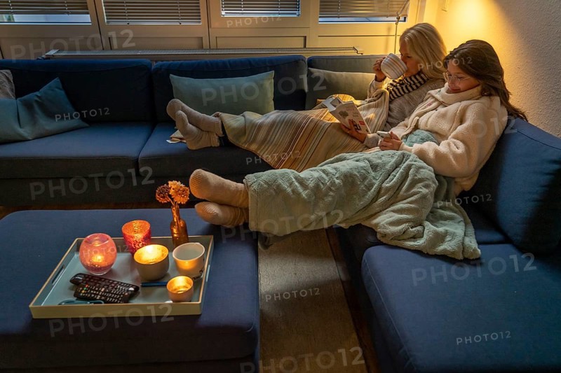 Symbolic image energy saving, cold winter, energy crisis, cold flat, mother and daughter lying on the sofa, warmly dressed, candlelight, heating on to a minimum
