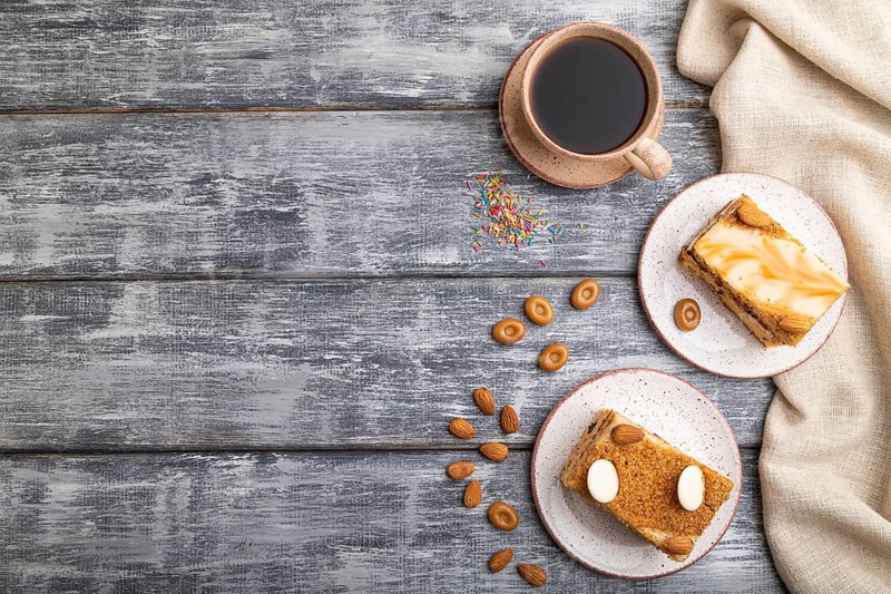 Honey cake with milk cream, caramel, almonds and a cup of coffee on a gray wooden background and linen textile. Top view, flat lay, copy space