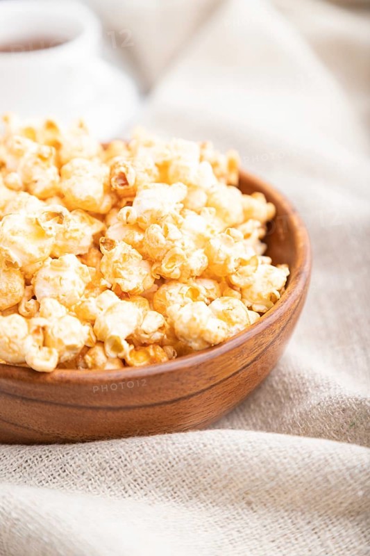 Popcorn with caramel in wooden bowl and a cup of coffee on a white wooden background and linen textile. Side view, close up, selective focus