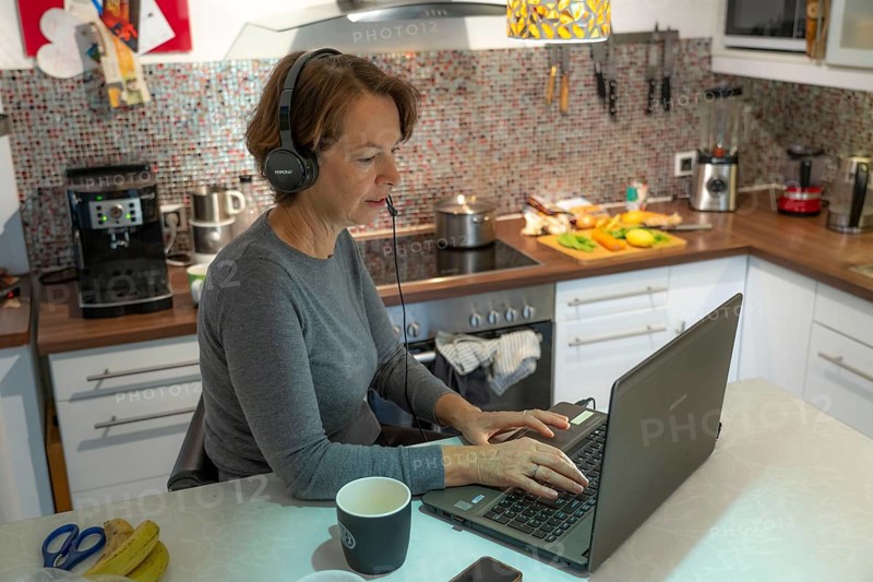 Woman, mid-50s, works from home, with laptop and communicates with colleagues via headset, home office, in the kitchen