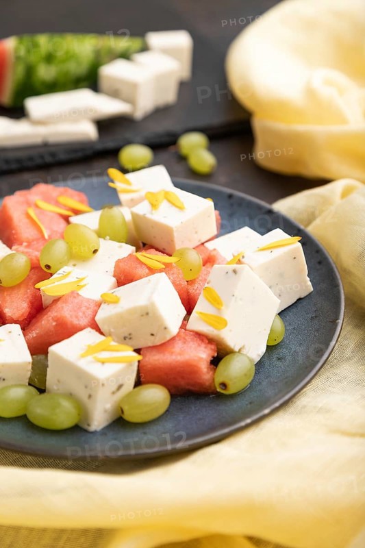 Vegetarian salad with watermelon, feta cheese, and grapes on blue ceramic plate on black concrete background and yellow linen textile. Side view, close up, selective focus