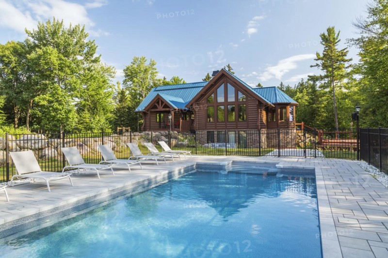 In-ground salt water swimming pool and brown stained milled Eastern white pine timber and flat log profile home facade with stone cladding on walk-out lower level and blue standing-seam sheet metal roof, Quebec, Canada, North America