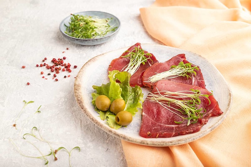 Slices of smoked salted meat with cilantro microgreen on gray concrete background and orange textile. Side view, close up