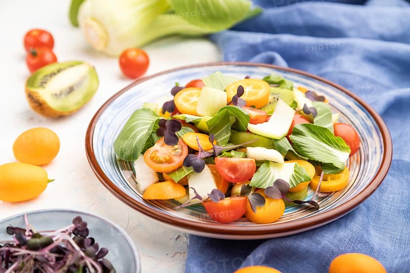 Vegetarian salad of pac choi cabbage, kiwi, tomatoes, kumquat, microgreen sprouts on a white concrete background and blue linen textile. Side view, close up, selective focus