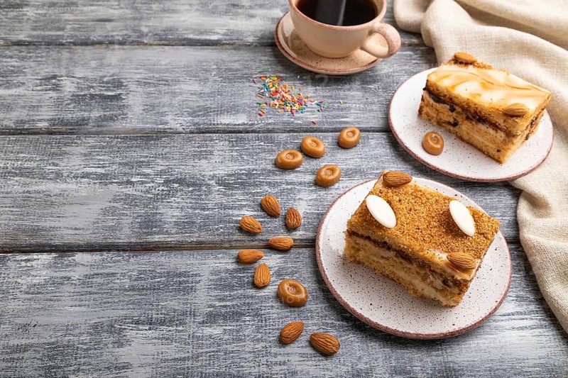 Honey cake with milk cream, caramel, almonds and a cup of coffee on a gray wooden background and linen textile. Side view, copy space