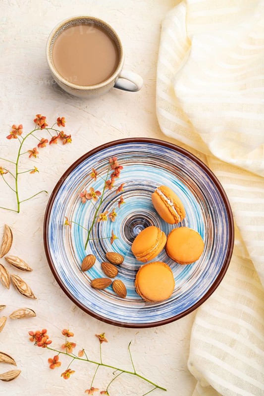 Orange macarons or macaroons cakes with cup of coffee on a white concrete background and linen textile. Top view, flat lay, close up