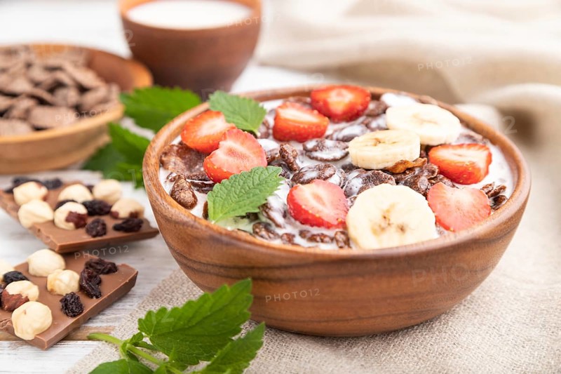Chocolate cornflakes with milk and strawberry in wooden bowl on white wooden background and linen textile. Side view, close up, selective focus