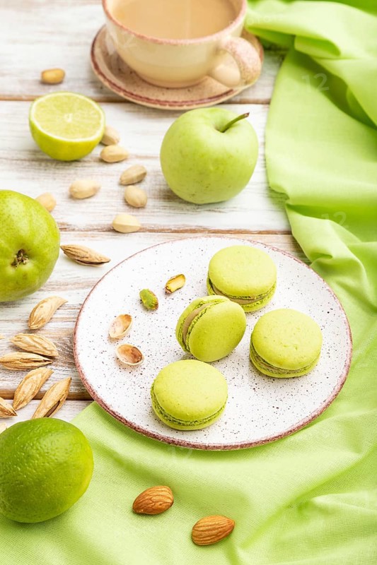 Green macarons or macaroons cakes with cup of coffee on a white wooden background and green linen textile. Side view, close up