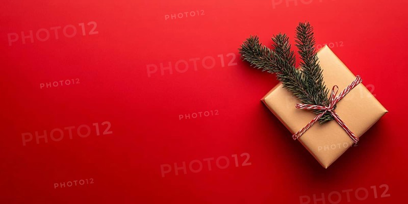 Banner with small Christmas gift box wrapped in graft paper with fir branch on side of red background with copy space. Generative Ai, AI generated