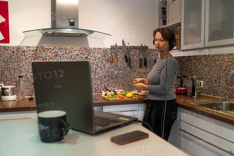 Woman, mid-50s, works from home, with laptop and communicates with colleagues via headset, home office, in the kitchen, while cooking