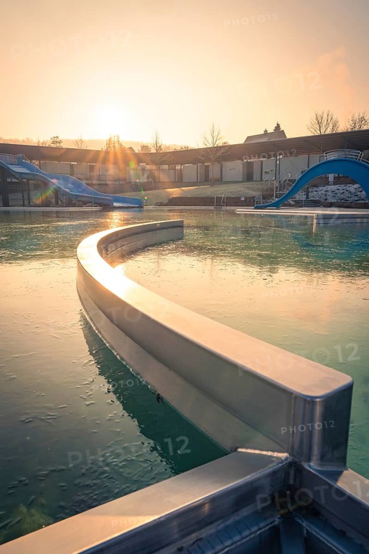 An outdoor pool with a curved water slide, flooded with warm morning sunshine and frozen water, ENCW outdoor pool Calw, Stammheim, Black Forest, Germany, Europe
