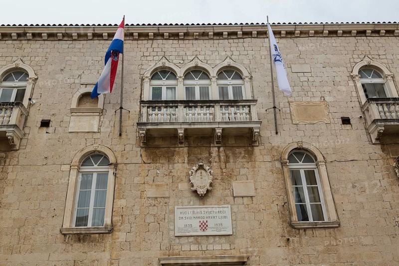 Old beige and tan stone cladded Trogir Town Hall building facade with balcony, flags, arched windows and commemorative 1835-1935 plaque, Trogir, Croatia, Europe