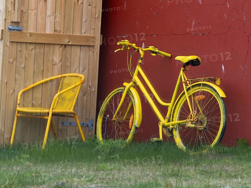 A bright yellow bicycle and matching chair placed outdoors by a wooden door and red wall, surrounded by grass, Yellow painted chair and bicycle, on a house building, in the town of Kiberg, Varanger Fjord, Finnish Lapland, Finland, Europe