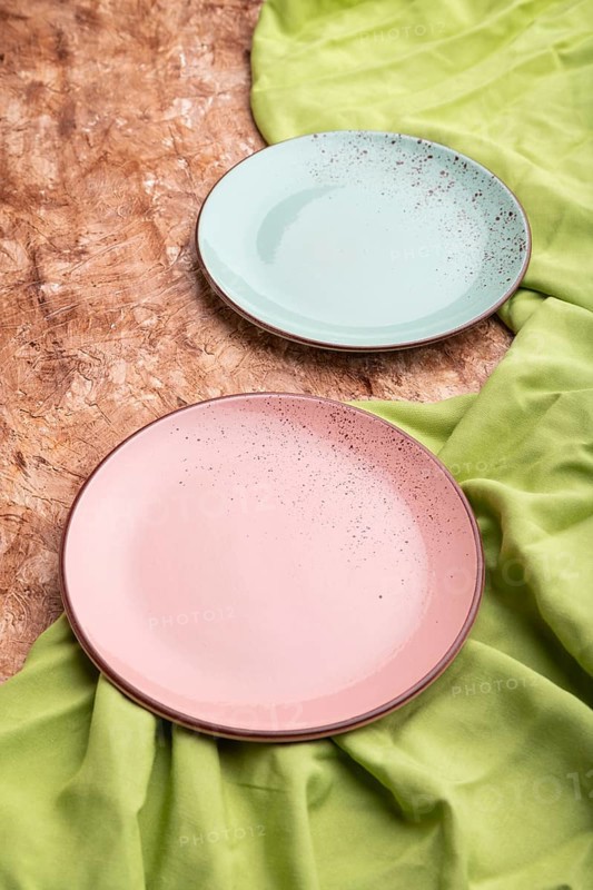 Two empty blue and pink plates on brown concrete background and green textile. Side view, close up