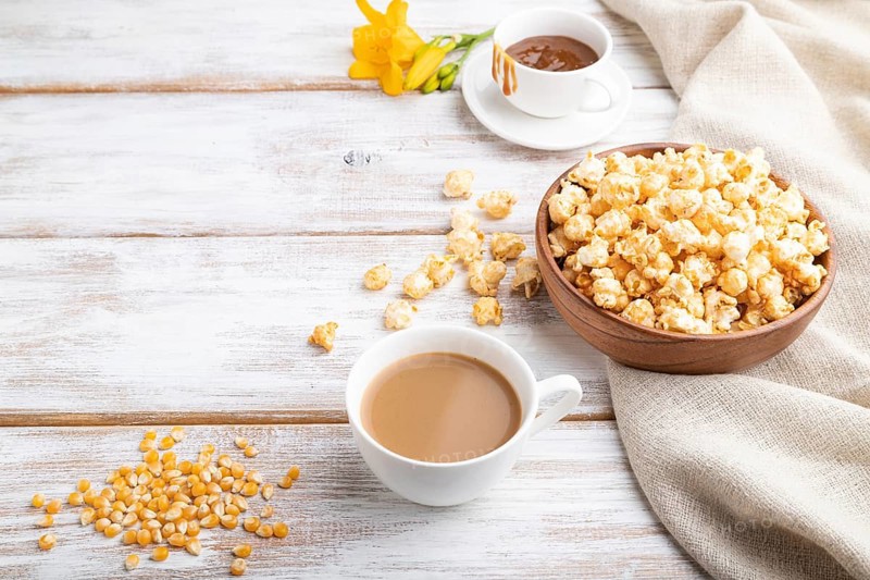 Popcorn with caramel in wooden bowl and a cup of coffee on a white wooden background and linen textile. Side view, copy space