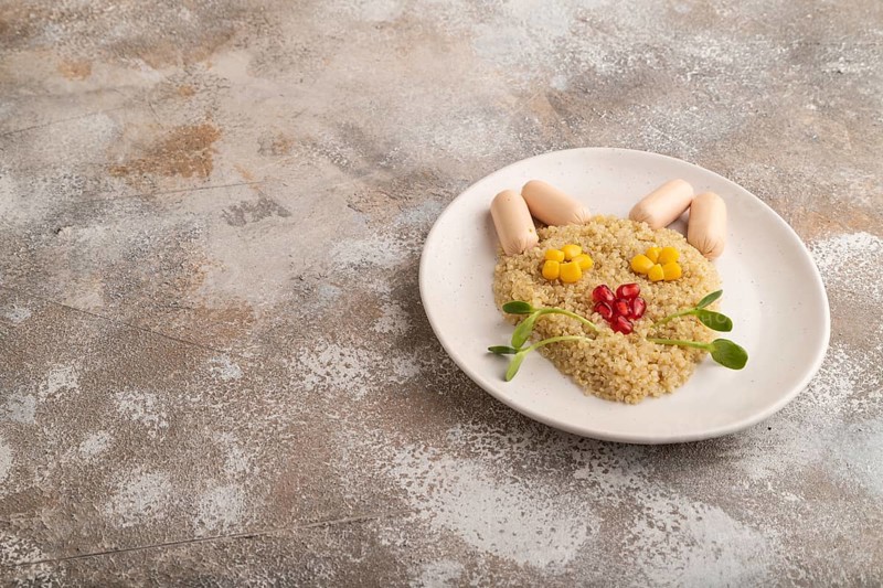 Funny mixed quinoa porridge, sweet corn, pomegranate seeds and small sausages in form of cat face on brown concrete background. Side view, copy space. Food for children, healthy food concept