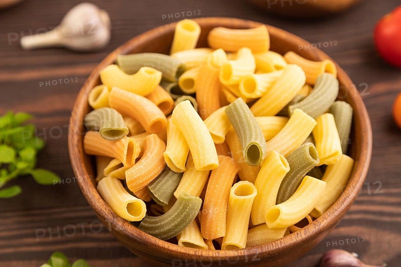 Rigatoni colored raw pasta with tomato, eggs, spices, herbs on brown wooden background. Side view, close up, selective focus