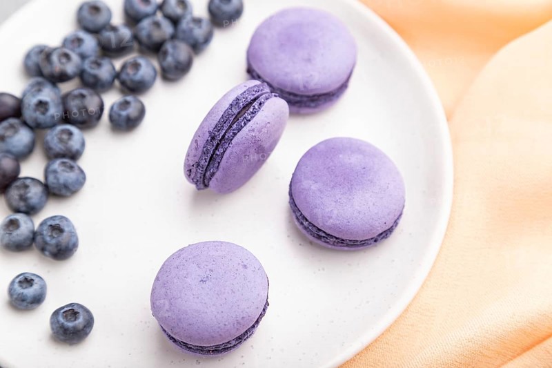 Purple macarons or macaroons cakes with blueberries on white ceramic plate on a gray concrete background and orange textile. Top view, close up, selective focus