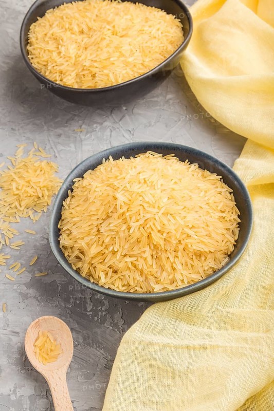 Blue ceramic bowl with raw golden rice and wooden spoon on a gray concrete background and yellow textile. Side view, close up