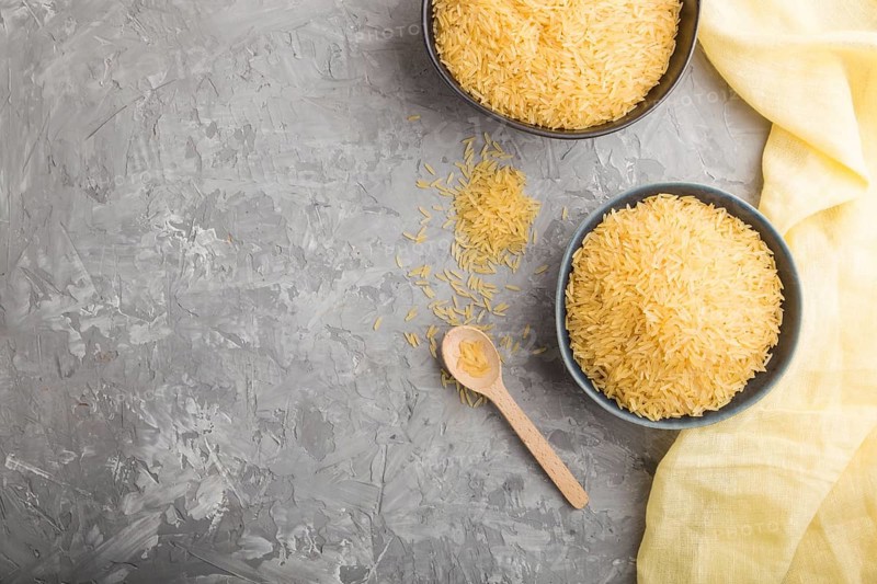 Blue ceramic bowl with raw golden rice and wooden spoon on a gray concrete background and yellow textile. Top view, flat lay, copy space