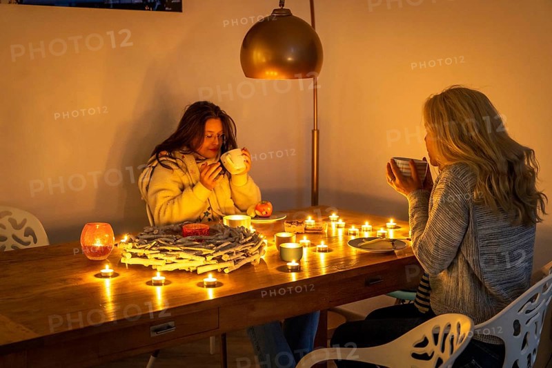 Symbolic image energy saving, cold winter, energy crisis, cold flat, mother and daughter dressed warmly at dinner, only by candlelight, heating on to a minimum