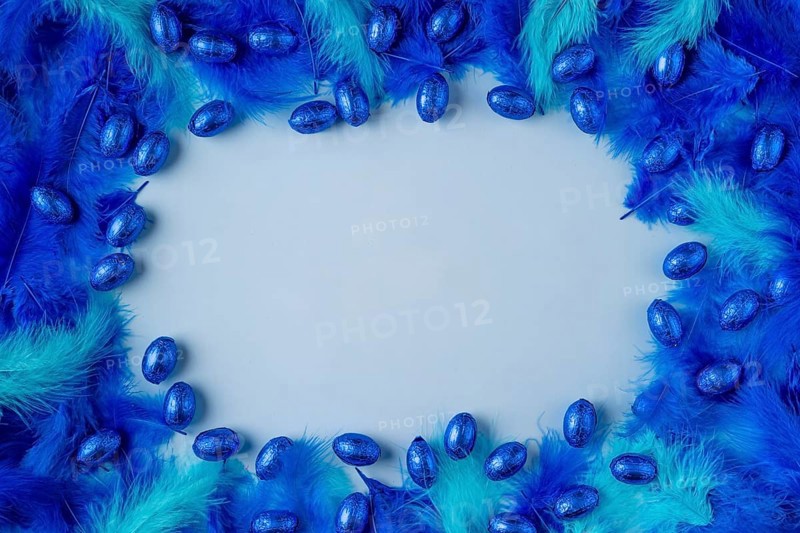 Frame made of blue and turquoise feathers with blue chocolate eggs on a light blue background