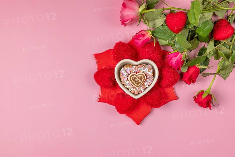 Bouquet of roses next to a heart-shaped bowl with a muffin surrounded by red hearts on a pink background