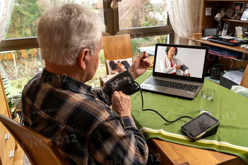 Symbolic image of telemedicine, elderly patient speaks to a doctor in a video conference from home, the use of a blood pressure monitor is explained