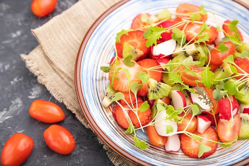 Vegetarian fruits and vegetables salad of strawberry, kiwi, tomatoes, microgreen sprouts on black concrete background and linen textile. Side view, close up, selective focus