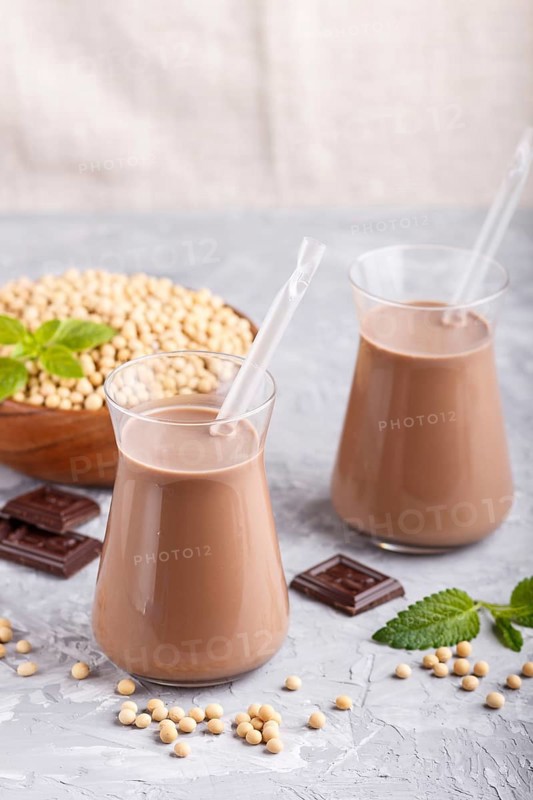 Organic non dairy soy chocolate milk in glass and wooden plate with soybeans on a gray concrete background. Vegan healthy food concept, close up, side view