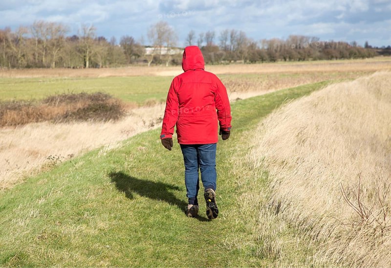 Woman wearing red winter jacket walking on raised coastal flood defence dyke embankment, Hollesley Bay, Suffolk, England, UK
