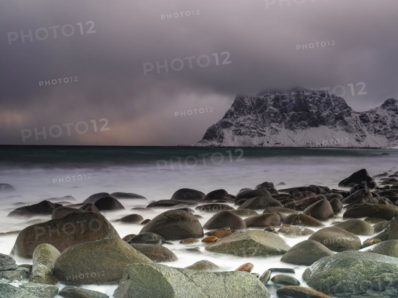 Rounded rocks on Utakleiv beach in a dramatic cloudy atmosphere, snow-capped mountains in the background, Vestvagoy Island, Lofoten, Norway, Europe