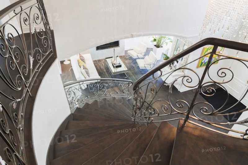 Looking down spiraling wooden staircase with wrought iron balusters onto entry way and living room with white leather sofa and armchairs, glass-top coffee table inside a luxurious residential home, Quebec, Canada, North America