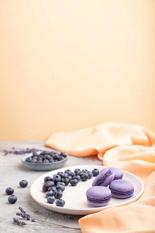 Purple macarons or macaroons cakes with blueberries on white ceramic plate on a gray and orange background and orange textile. Side view, copy space, selective focus