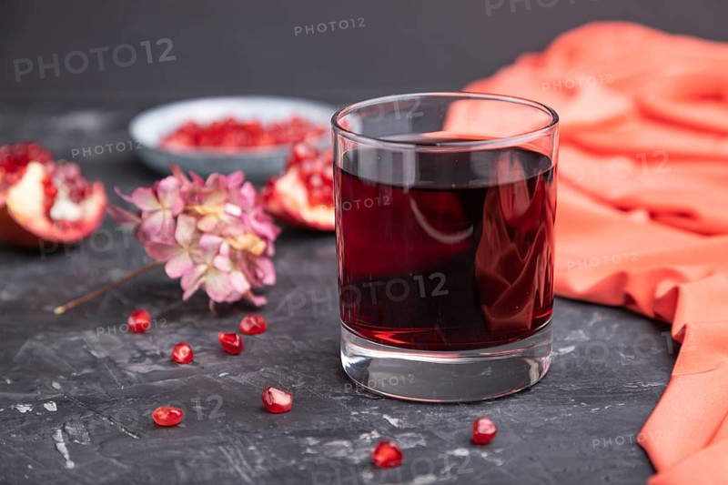 Glass of pomegranate juice on a black concrete background with red textile. Side view, close up, selective focus
