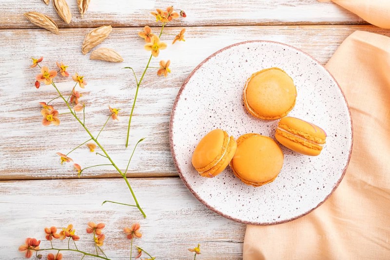 Orange macarons or macaroons cakes with cup of apricot juice on a white wooden background and orange linen textile. Top view, flat lay, close up