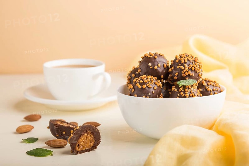 Chocolate caramel ball candies with almonds and a cup of coffee on a white and orange background and yellow textile. Side view, close up, selective focus