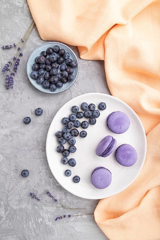 Purple macarons or macaroons cakes with blueberries on white ceramic plate on a gray concrete background and orange textile. Flat lay, top view, close up