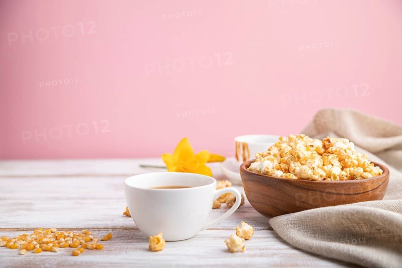 Popcorn with caramel in wooden bowl and a cup of coffee on a white and pink background and linen textile. Side view, copy space, selective focus