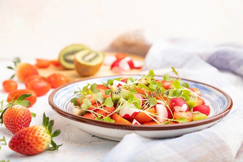 Vegetarian fruits and vegetables salad of strawberry, kiwi, tomatoes, microgreen sprouts on white concrete background and linen textile. Side view, close up, selective focus