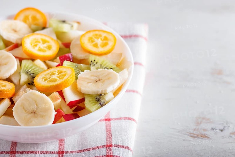 Vegetarian salad of bananas, apples, pears, kumquats and kiwi on linen tablecloth, close up, selective focus, copy space. white wooden background