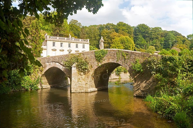 Bridge crossing the River Frome and the classical Georgian facade of Iford Manor, near Freshford, Wiltshire, England, UK