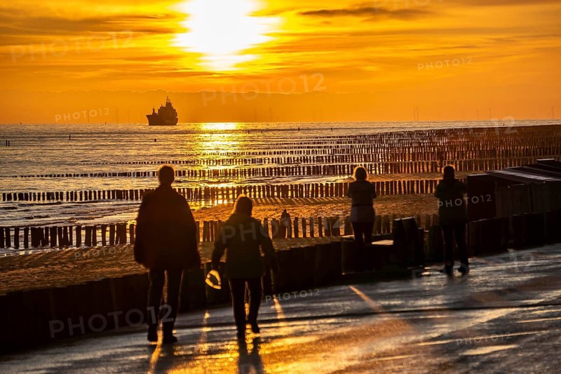 Sunset on the beach of Zoutelande, beach with wooden pile breakwaters, tourists, cargo ship sailing towards the Westerschelde, Zeeland, Netherlands