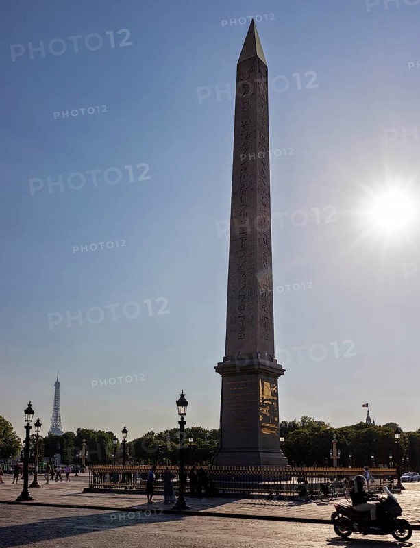 Sunbeams behind an obelisk, Eiffel Tower in the background, a motorbike passing by, Paris