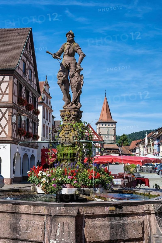 The market square fountain, Röhrbrunnen, with a knight on the shield the city coat of arms, behind it the Obertorturm, Haigeracher Tor, historic town tower and landmark in the old town centre of Gengenbach, Ortenaukreis, Baden-Württemberg, Germany, Europe