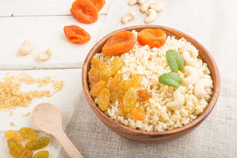 Bulgur porridge with dried apricots, raisins and cashew in wooden bowl on a white wooden background and linen textile. Side view, close up. Turkish traditional cuisine