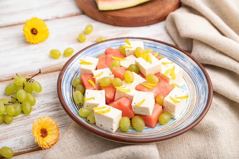 Vegetarian salad with watermelon, feta cheese, and grapes on blue ceramic plate on white wooden background and linen textile. Side view, close up, selective focus